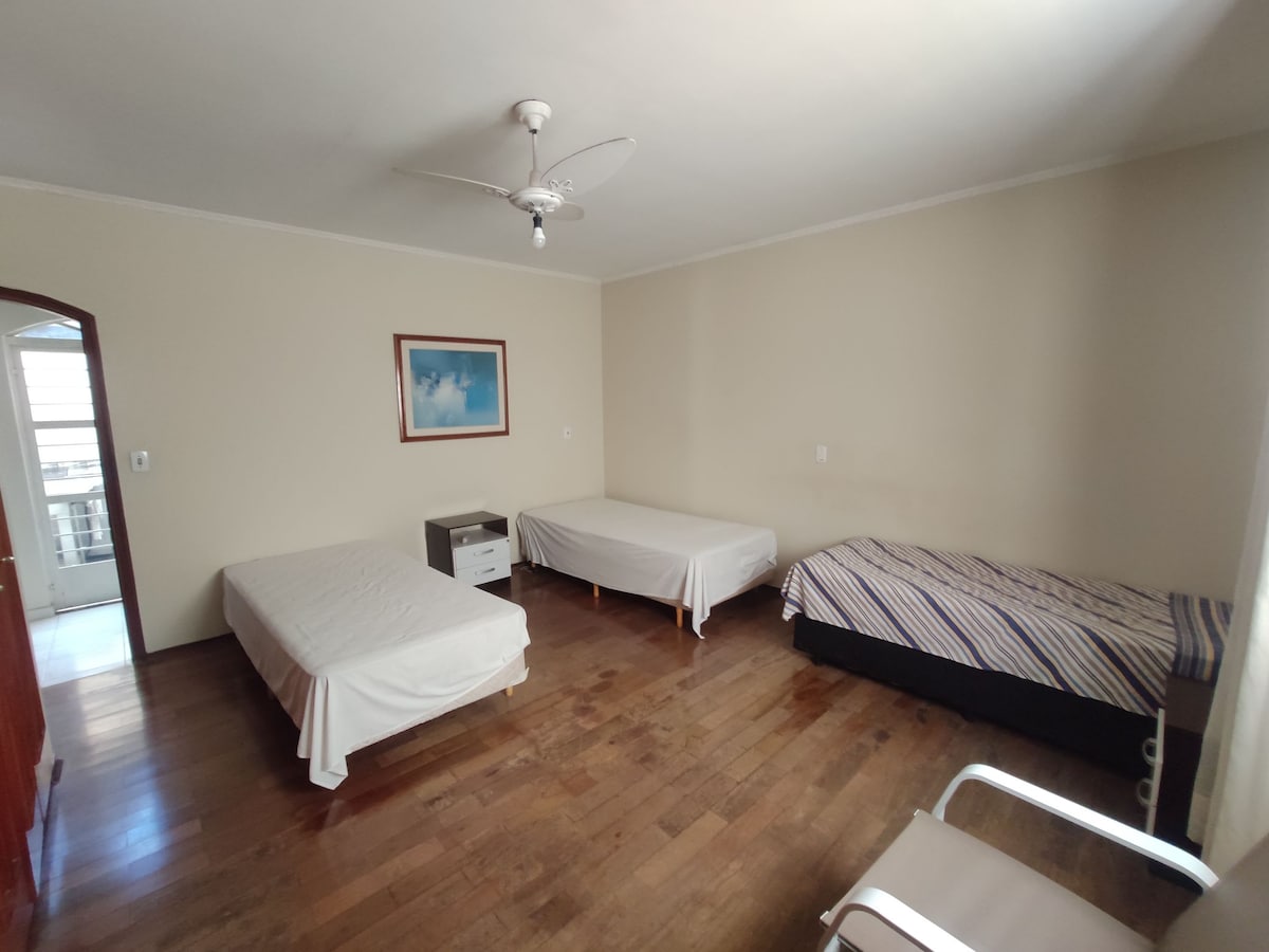 A spacious bedroom features three beds, including a striped twin-size bed and two simple twin beds. A small nightstand is positioned beside one bed. Light wood flooring complements the neutral-colored walls, while a ceiling fan is centrally located above.