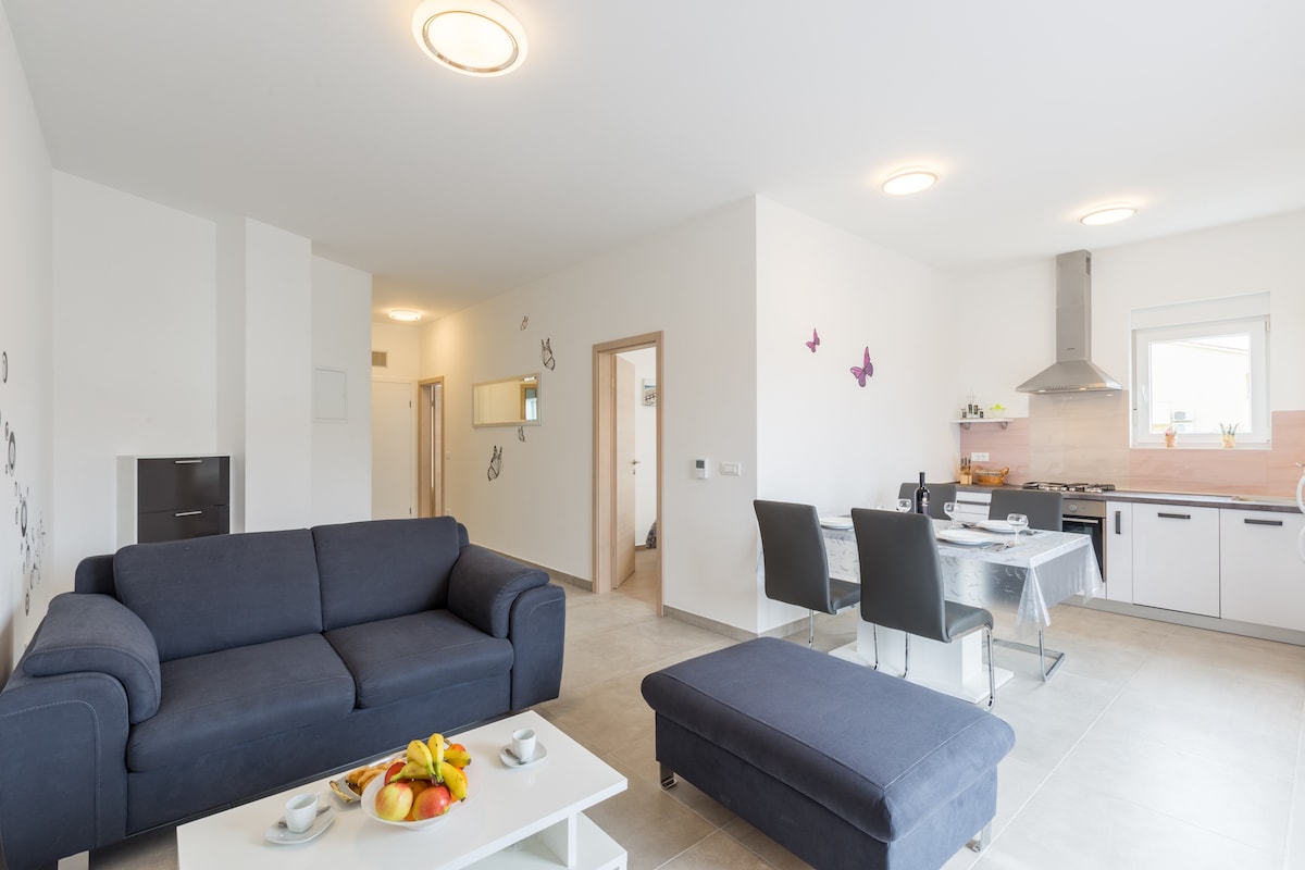 A modern living area features a dark blue sofa and a matching ottoman, with a white coffee table displaying fresh fruit. The kitchen area, visible in the background, includes a dining table set for meals and light-colored cabinetry. Soft lighting enhances the neutral decor.