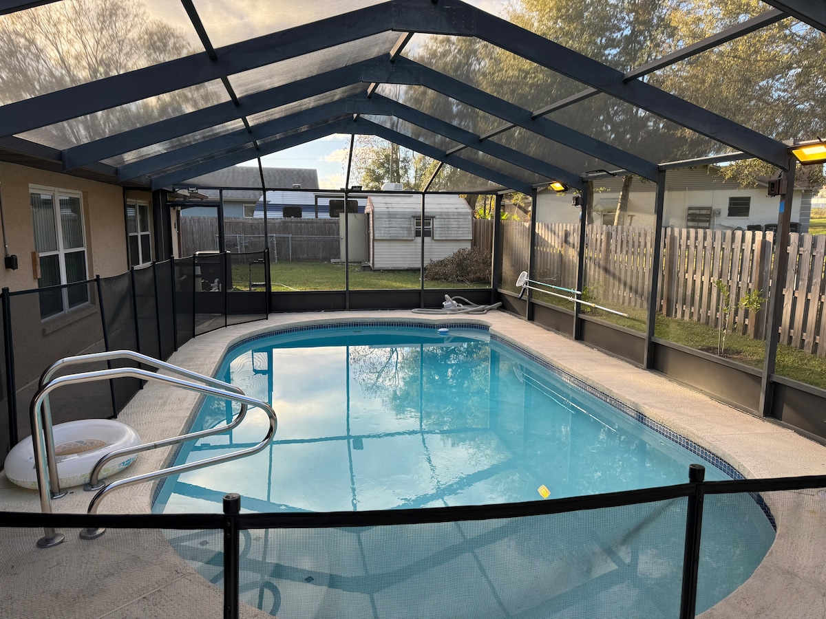 A screened-in pool area features a clear blue pool surrounded by safety fencing. Overhead lighting illuminates the space, and a gentle slope leads to the pool steps. The yard beyond offers privacy with fencing and greenery visible in the background.