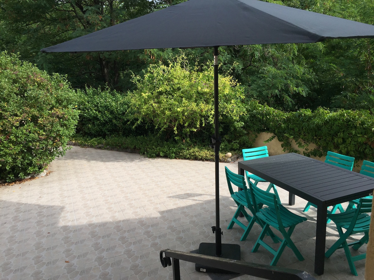An outdoor terrace features a large, dark umbrella providing shade over a rectangular table surrounded by turquoise chairs. The area is framed by lush greenery, creating a peaceful environment for relaxation or dining.