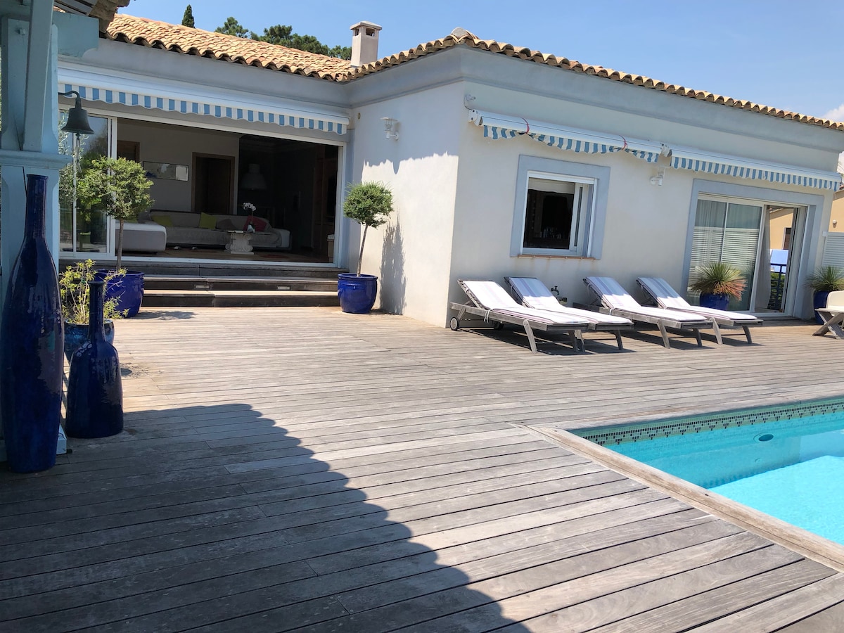 The image displays a sunlit wooden deck beside a pool. Lounge chairs are neatly arranged, and vibrant blue pots are positioned along the sides. The entrance to a spacious living area is visible, featuring large glass doors that open onto the deck.