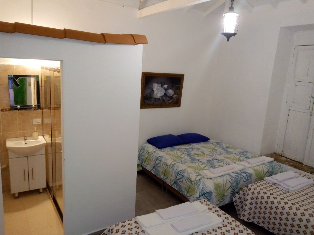 A spacious room is presented with a large king-size bed and two double beds, each featuring fresh linens. An adjoining bathroom with a transparent shower and sink is visible. Soft lighting from a hanging fixture enhances the bright and airy feel of the space.