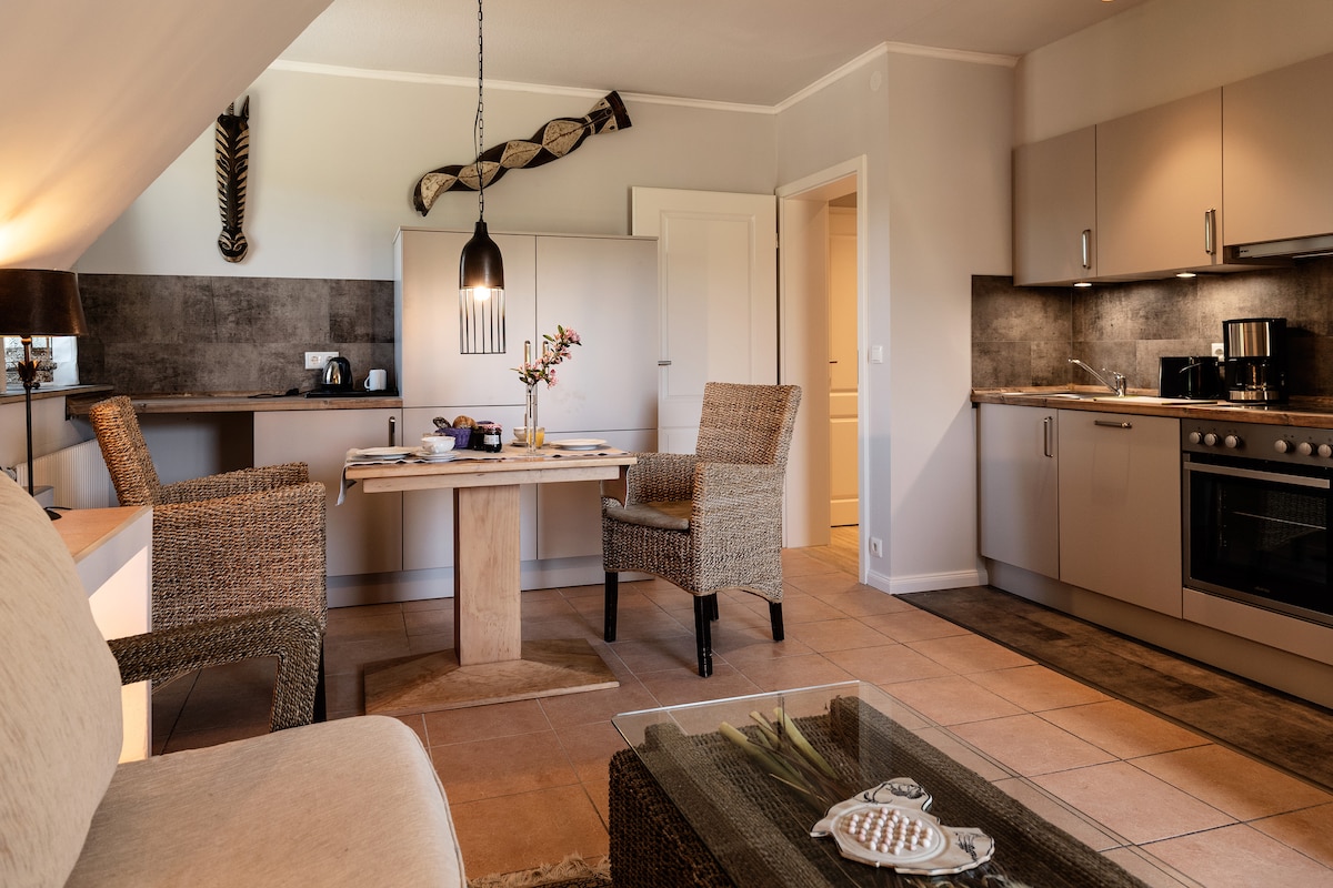 A dining area is set with a wooden table surrounded by two chairs, positioned near a modern kitchen with sleek cabinetry. A light fixture hangs above the table, adding an element of style. The open layout enhances the spacious feel of the living area.