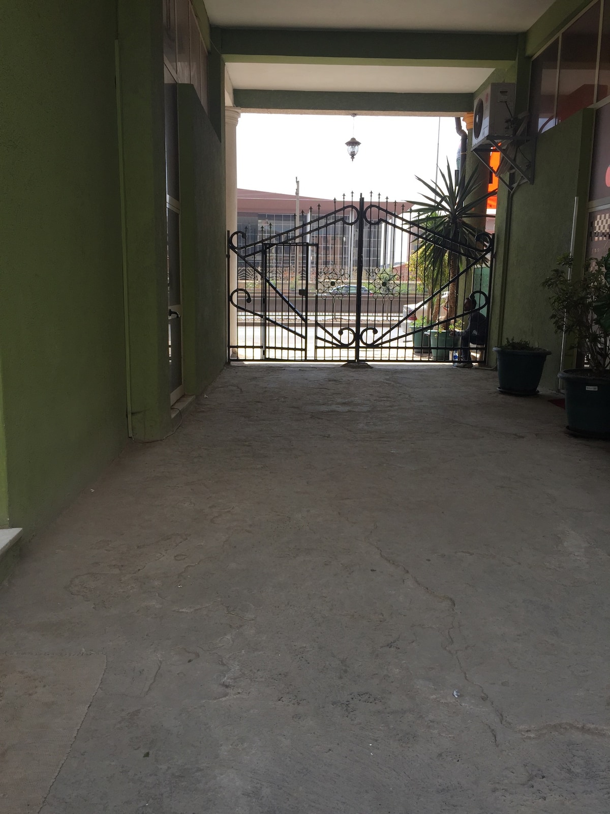 An open corridor is seen leading to an iron gate, framed by green walls. Potted plants are positioned along the sides, providing a touch of greenery. Natural light filters through the entrance, illuminating the smooth, gray surface of the floor.