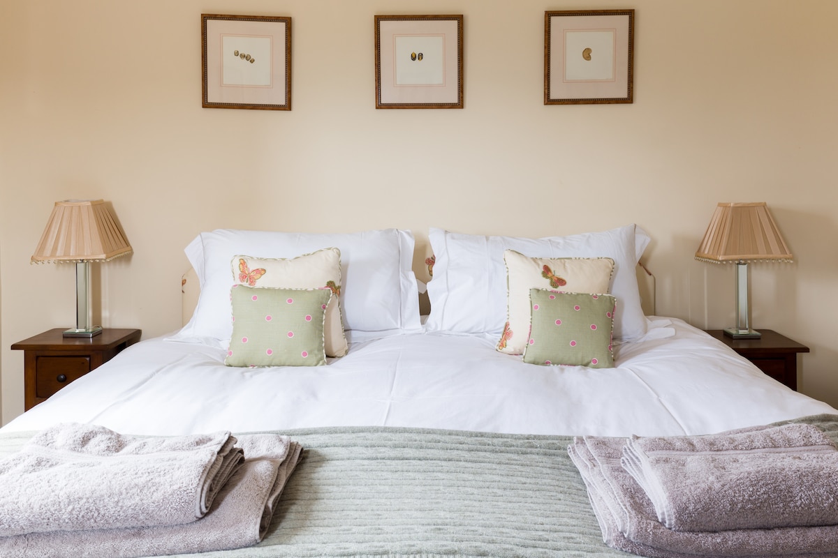A spacious bedroom features a super-king-sized bed adorned with white linens and decorative cushions. Two bedside lamps provide soft lighting, while framed artwork hangs above the bed. Folded towels are neatly arranged at the foot of the bed on a textured green blanket.