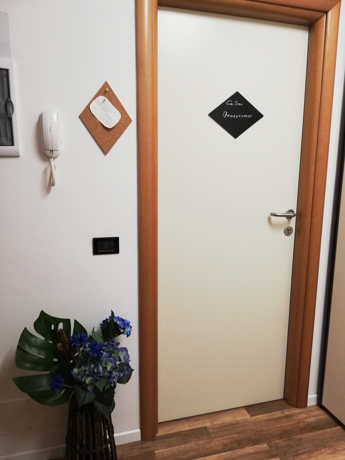 A neutral-toned door is framed by wooden trim, with a small decorative plaque labeled 'Guest Room'. A telephone and light switch are mounted nearby. A woven basket contains greenery and flowers, adding a touch of nature to the space.
