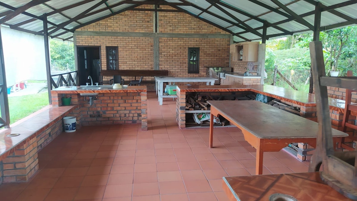 An open kitchen area is presented featuring brick walls and a tiled floor. Multiple countertops are available, along with a sink and stove. Wooden shelves store utensils, while logs are neatly arranged nearby. Natural light fills the space, enhancing the functional layout.