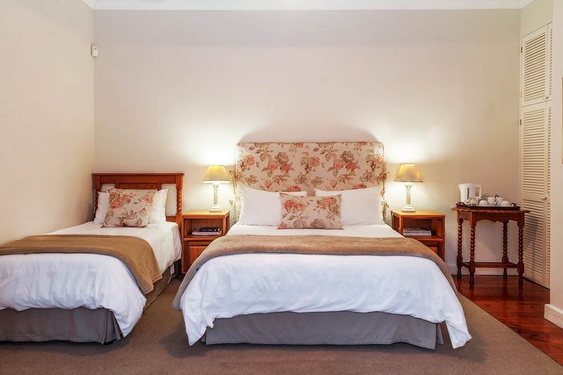 A well-appointed guest room features a spacious double bed and a single bed, adorned with floral-patterned headboards. Bedside tables with lamps provide soft lighting. A small table and tea/coffee-making facilities are visible against one wall, contributing to a comfortable and convenient atmosphere.