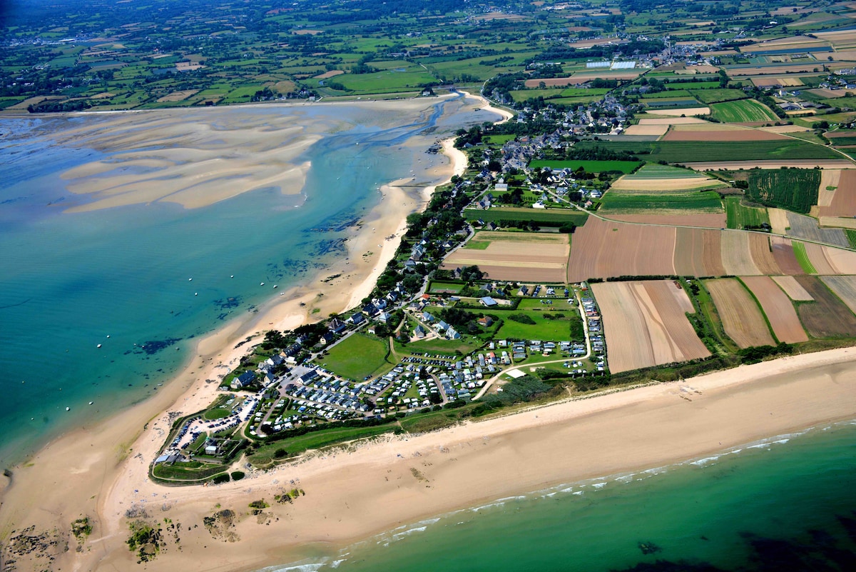 Aerial view captures the sandy beach gently meeting the coastline, bordered by agricultural fields and patches of greenery. The layout of the residential area is visible, along with the shoreline's curve leading to the distant village and water dotted with small boats.