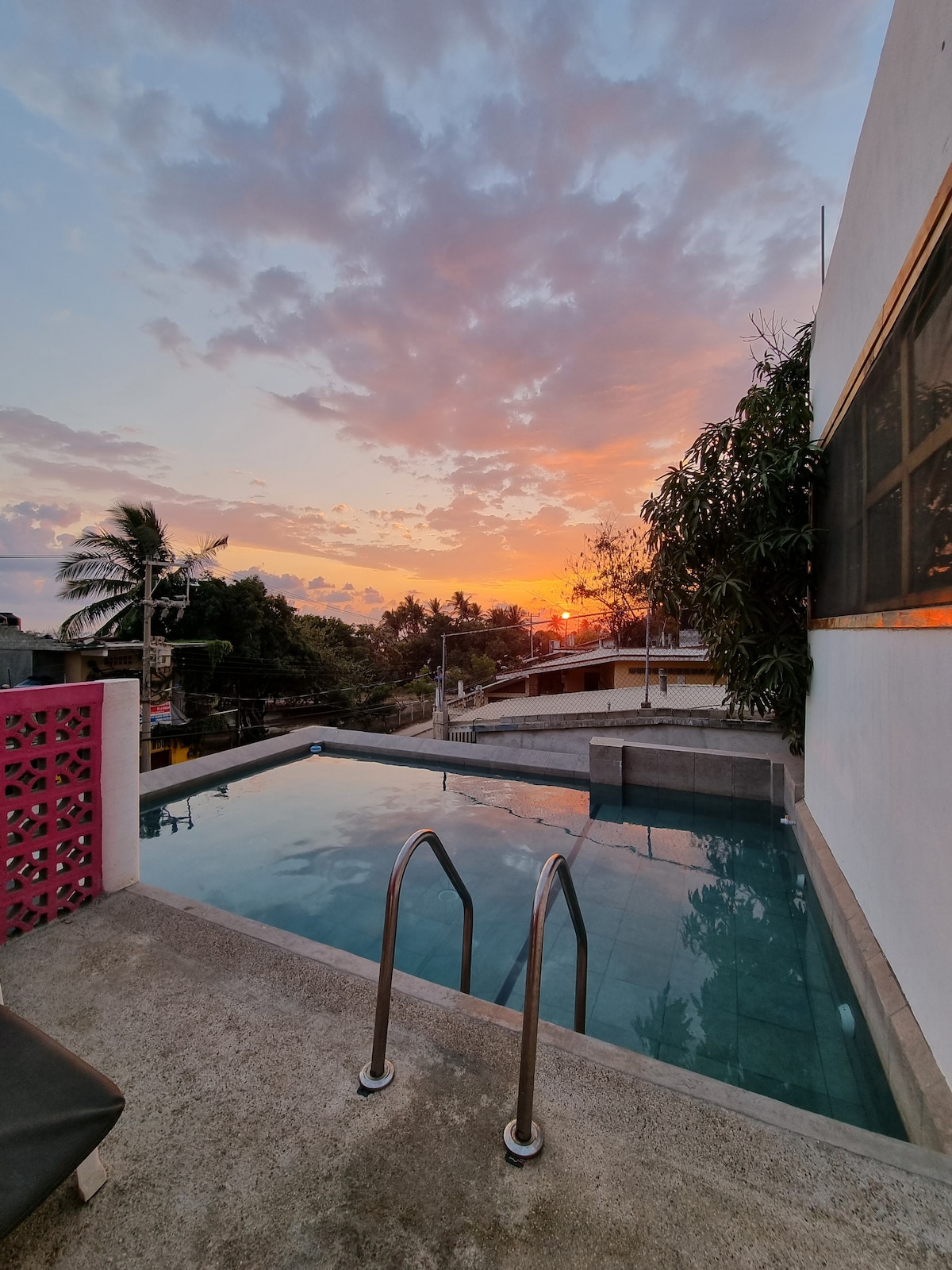 A refreshing pool is visible against a vibrant sunset sky, with clouds reflecting hues of orange and pink. Concrete steps lead into the pool, while tropical plants frame the scene, emphasizing the inviting atmosphere of relaxation.
