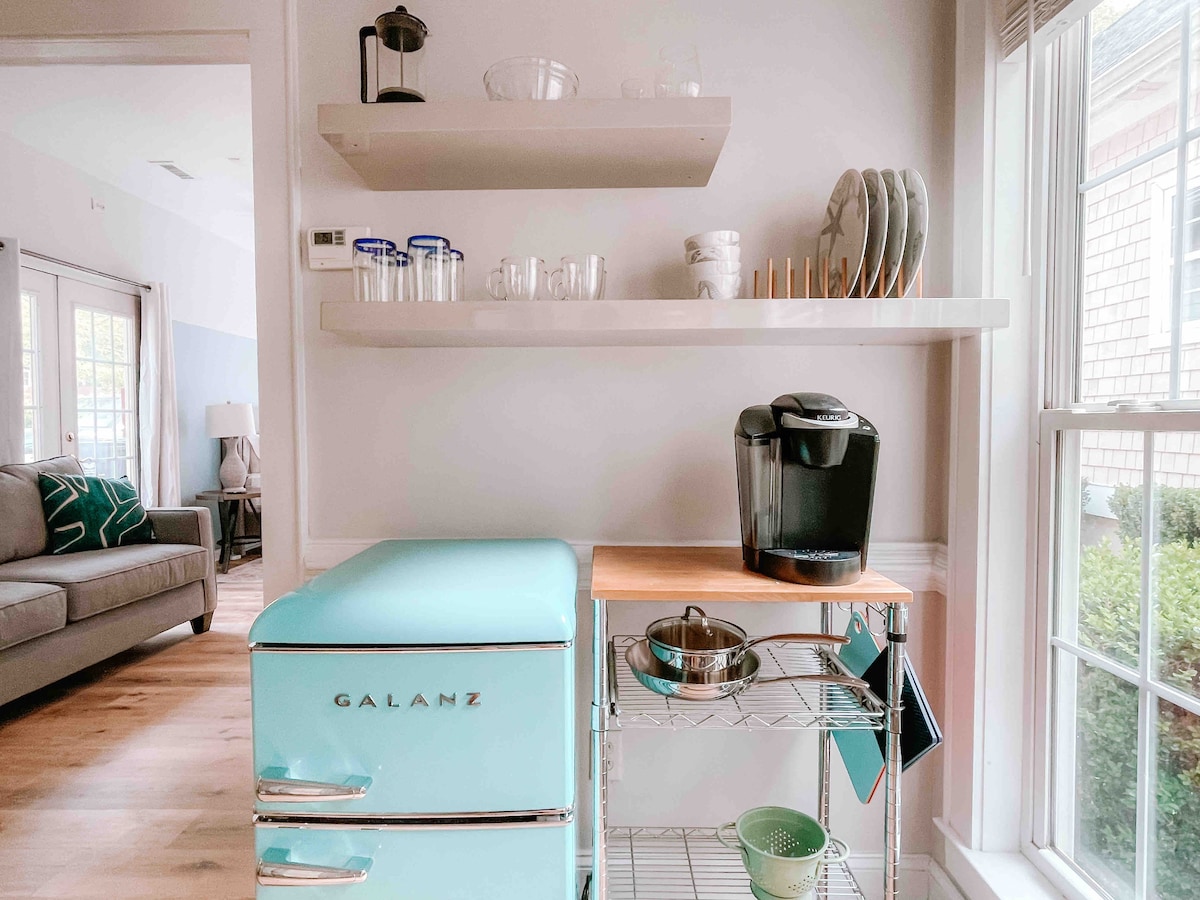 A retro-style refrigerator in a soft blue hue is positioned next to a wooden countertop. Above, open shelving displays glassware and dishware, while a coffee maker sits nearby. Natural light filters through a window, illuminating the organized and functional kitchen space.