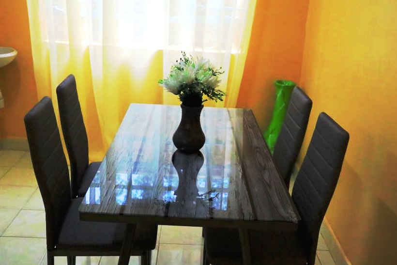 A dining area features a wooden table at its center, surrounded by four upholstered chairs. A vase filled with artificial flowers sits atop the table, offering a touch of decor. Natural light filters in through sheer curtains, softly illuminating the warm orange walls.