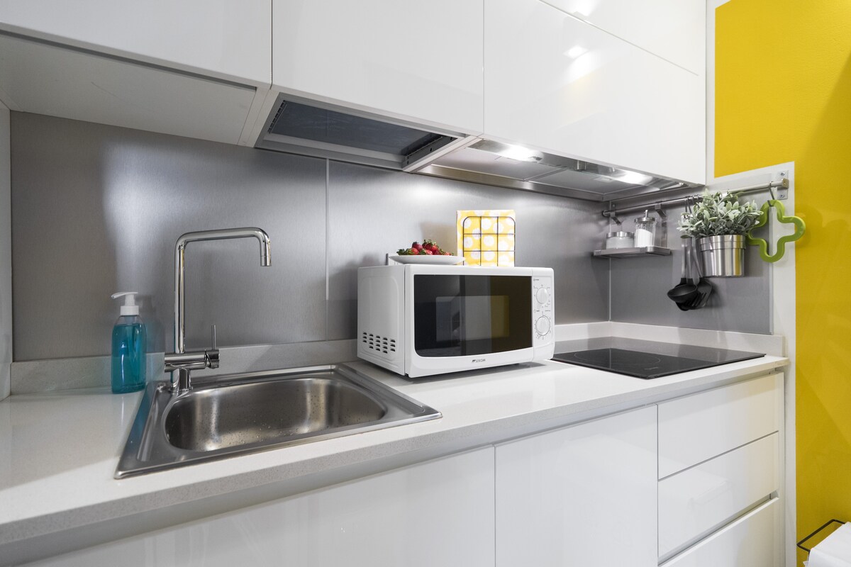 The kitchen features sleek white cabinetry and a modern countertop. Essential appliances include a microwave and an induction cooktop, complemented by a stainless steel sink. A small shelf holds potted plants, and the wall is decorated with vibrant yellow accents.