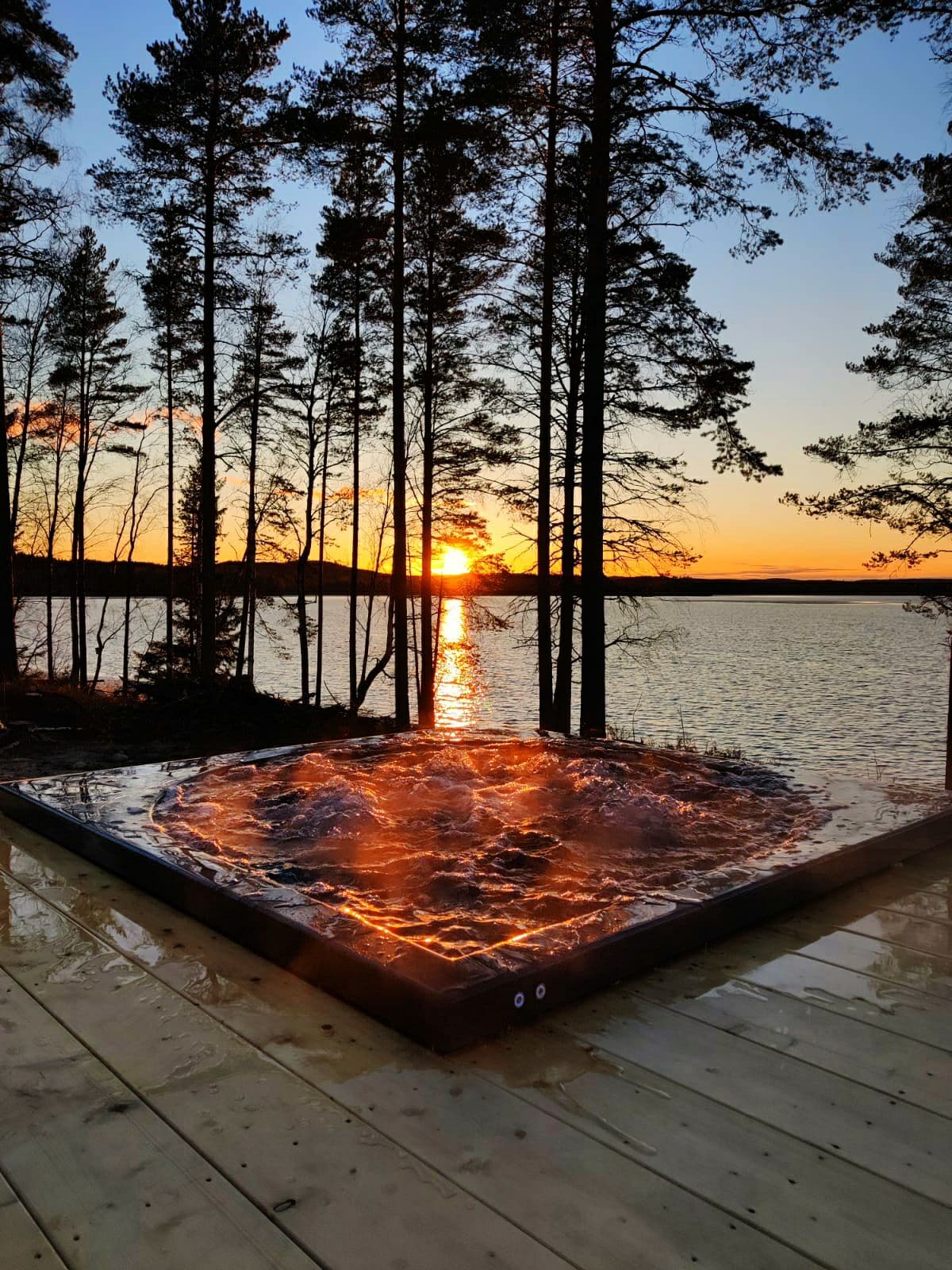 A hot tub is positioned on a wooden deck, surrounded by tall trees, with a stunning sunset reflecting off the water's surface. The golden hues contrast against the serene lake, creating a tranquil atmosphere for relaxation.