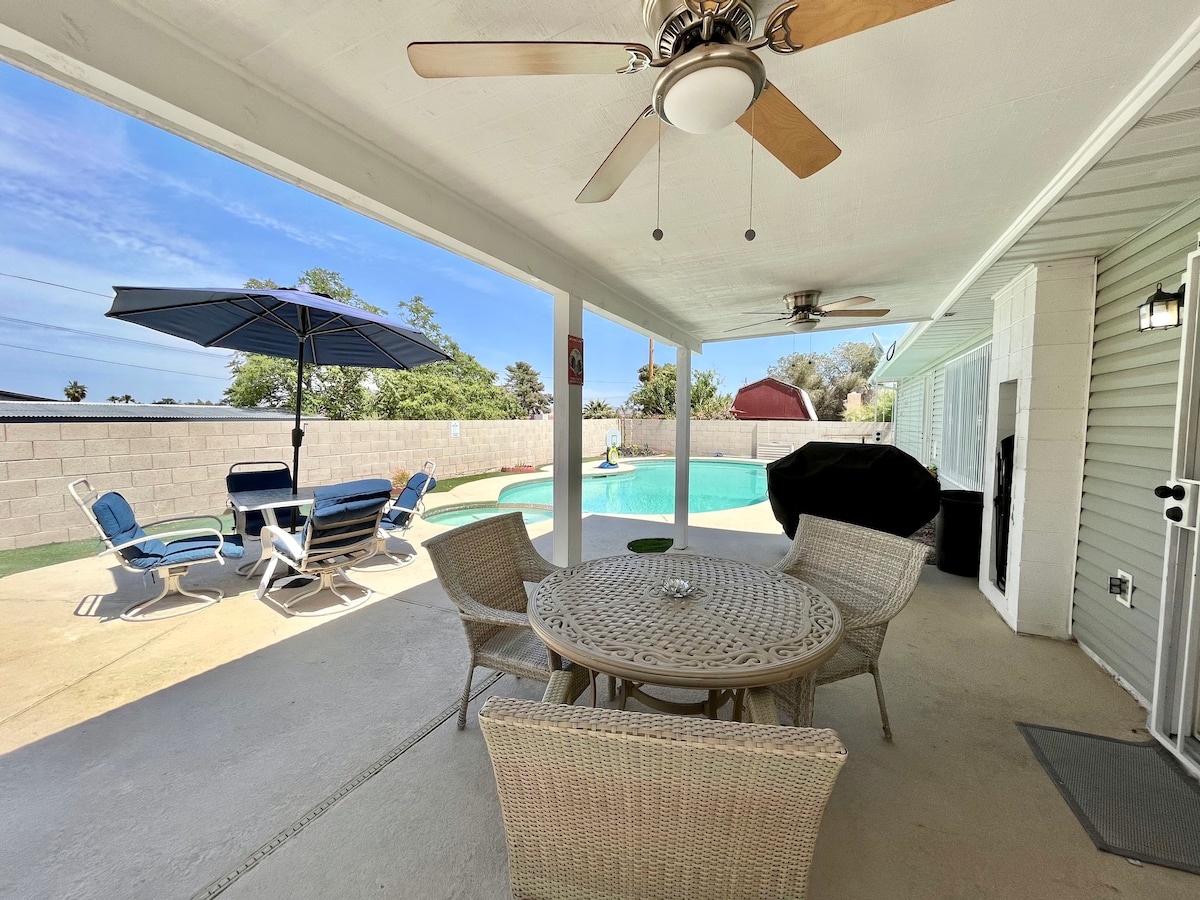 A shaded patio area is present, featuring a round wicker dining table surrounded by four chairs. A large umbrella offers protection from the sun, while a barbecue grill is positioned nearby. In the background, a swimming pool glistens under the clear sky.