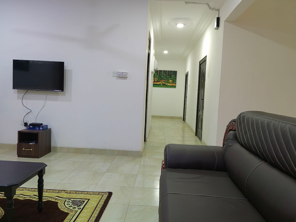 A spacious living area features a flat-screen TV mounted on the wall, with a dark coffee table situated on a decorative rug. A comfortable leather sofa is positioned to the right. The hallway in the background leads to additional rooms, with well-lit and neutral-colored walls.