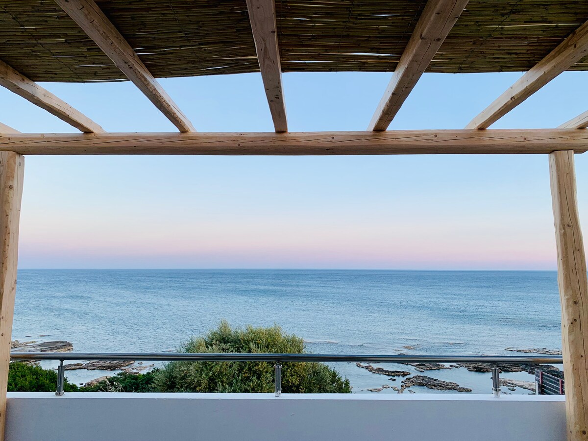 An ocean view is framed by wooden beams, showcasing a vast expanse of calm sea. The horizon is marked by soft pastel hues of blue and pink. Lush greenery is visible on the shoreline, enhancing the serene atmosphere.