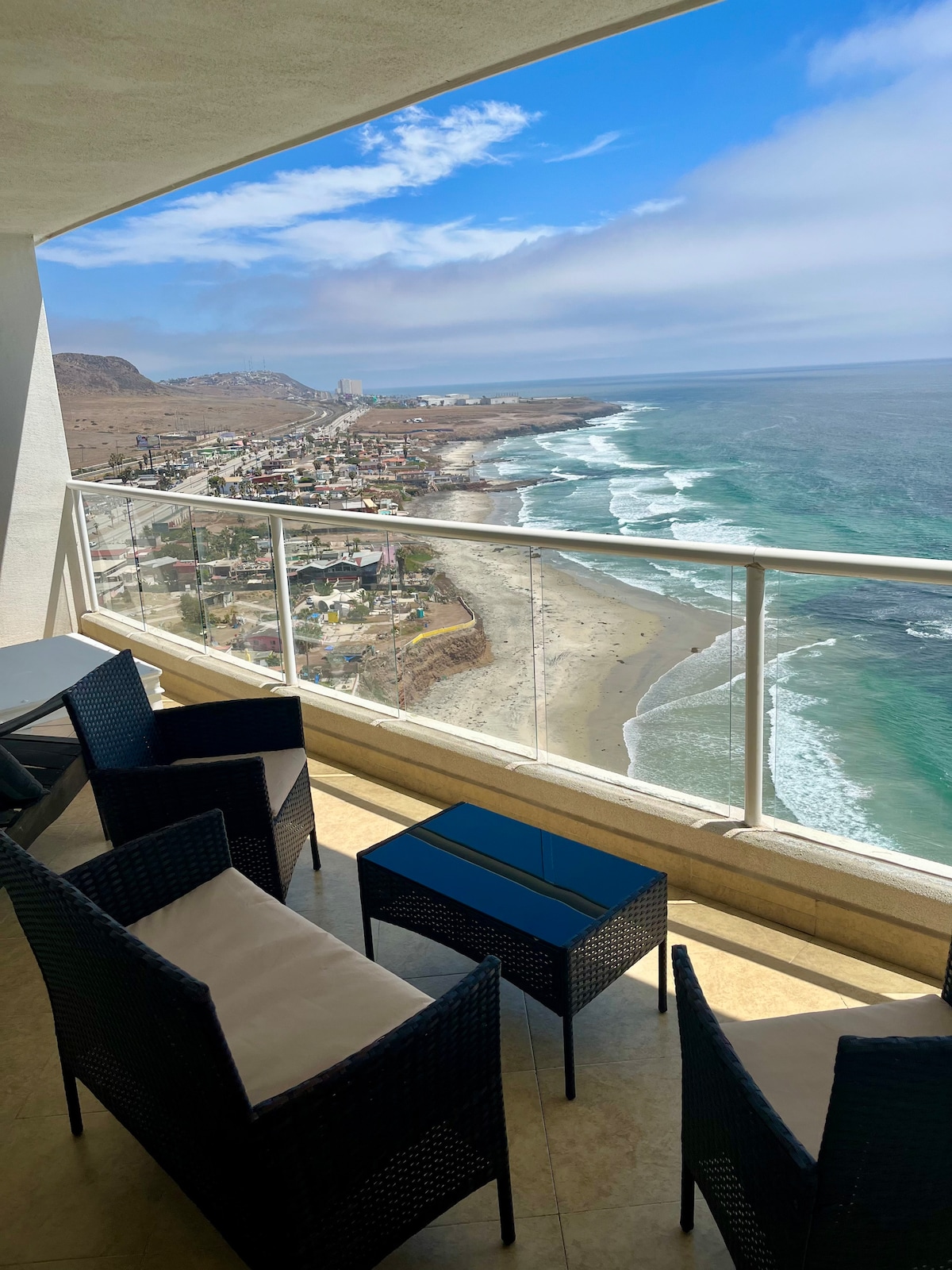 A balcony provides expansive views of the coastline and ocean waves. Four wicker chairs with cream-colored cushions surround a small black table, creating a relaxed seating area. The backdrop features a sandy beach and an undulating shoreline beneath a partly cloudy sky.