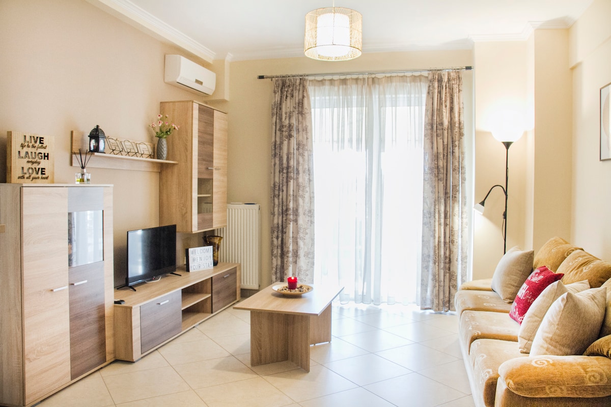 A bright living area features a cozy sofa adorned with decorative pillows. A wooden coffee table sits in the center, complemented by a television on a simple wooden stand. Floor-to-ceiling curtains frame a large window, allowing natural light to fill the space.