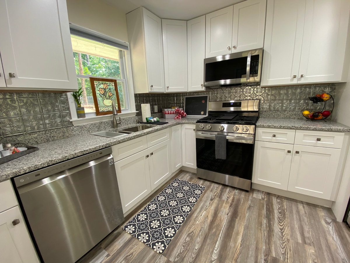 The kitchen features modern white cabinetry and granite countertops, complemented by a patterned rug. Stainless steel appliances include a refrigerator, dishwasher, and an oven with a microwave above. A window allows natural light, while decorative fruits are arranged on the counter.