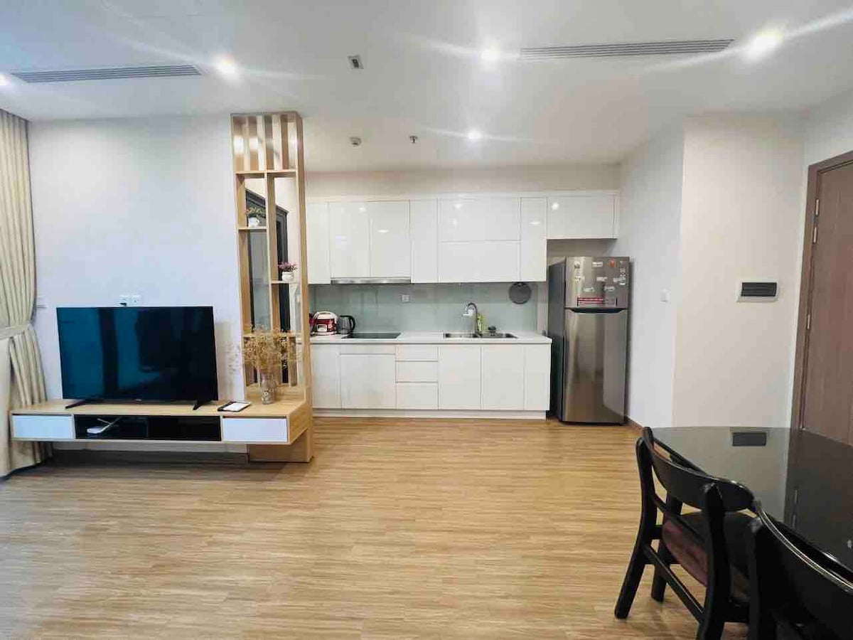 A modern kitchen space is featured with sleek white cabinetry and stainless steel appliances. A dining table with black chairs is positioned beside the kitchen, while a large television is visible in the living area. Natural light is provided by nearby windows.