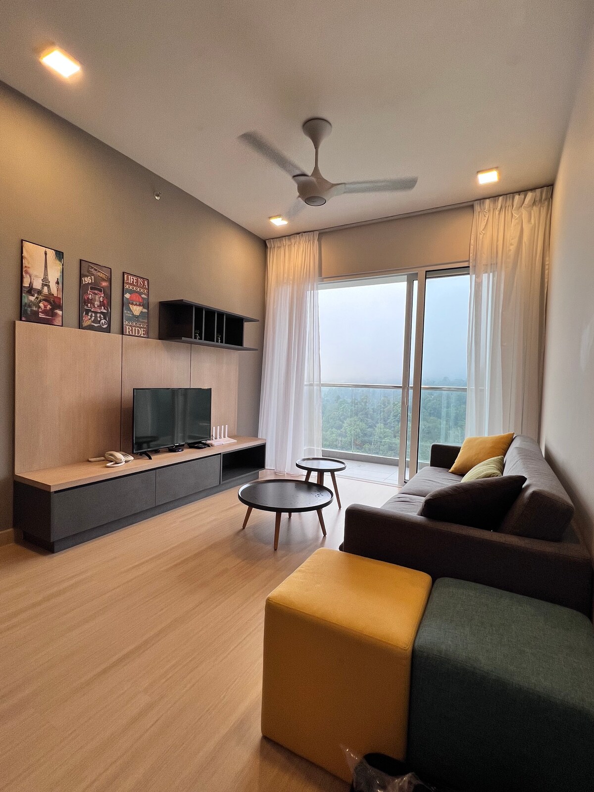 A cozy living area features a gray sofa complemented by a two-tone ottoman. A minimalist coffee table is positioned in front of a low entertainment unit, consisting of a television and decorative items. Large windows enhance the space, offering views of surrounding greenery.