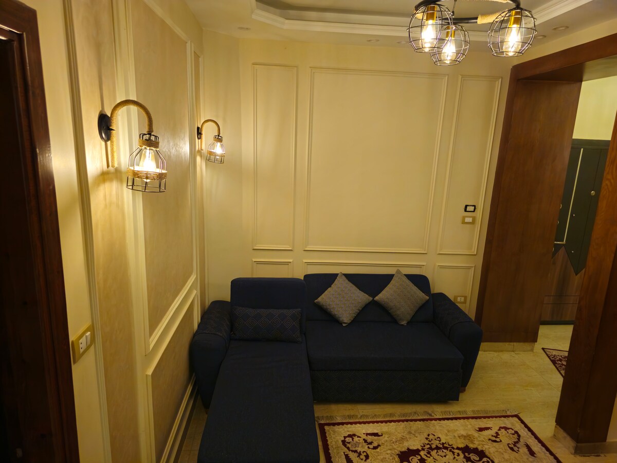 A cozy living area presents a modern blue sofa adorned with decorative cushions. Warm lighting is provided by stylish wall sconces, creating a comfortable ambiance. The soft carpet adds a touch of comfort to the space, enhancing the overall inviting atmosphere.