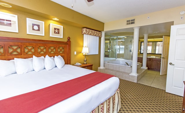 A spacious bedroom features a king bed adorned with white pillows and a burgundy bed runner. Decorative art pieces hang above the headboard. An en-suite area displays a jetted tub beside stone columns, while a bathroom with dual sinks is visible in the background.