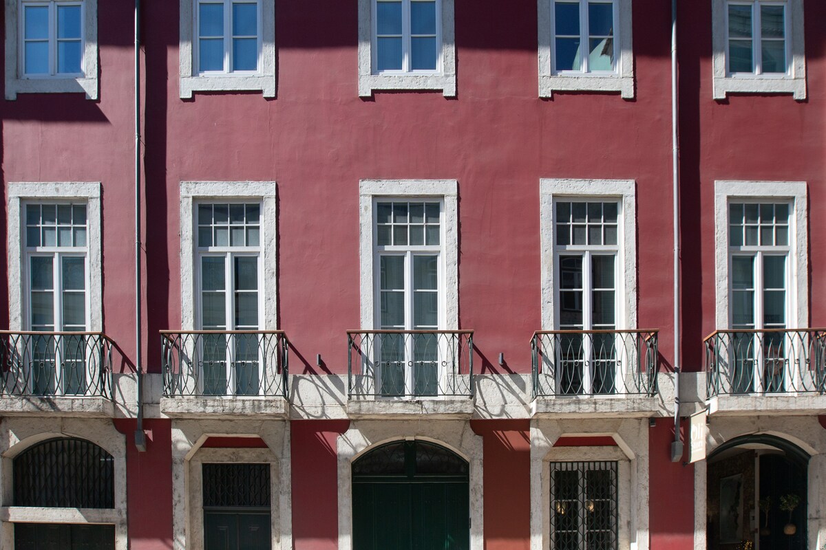 The historic 18th-century building features a vibrant pink facade with white trim. Four large windows, each adorned with decorative wrought iron balconies, provide ample natural light. The entrance showcases a green door, framed by symmetrical architectural details.