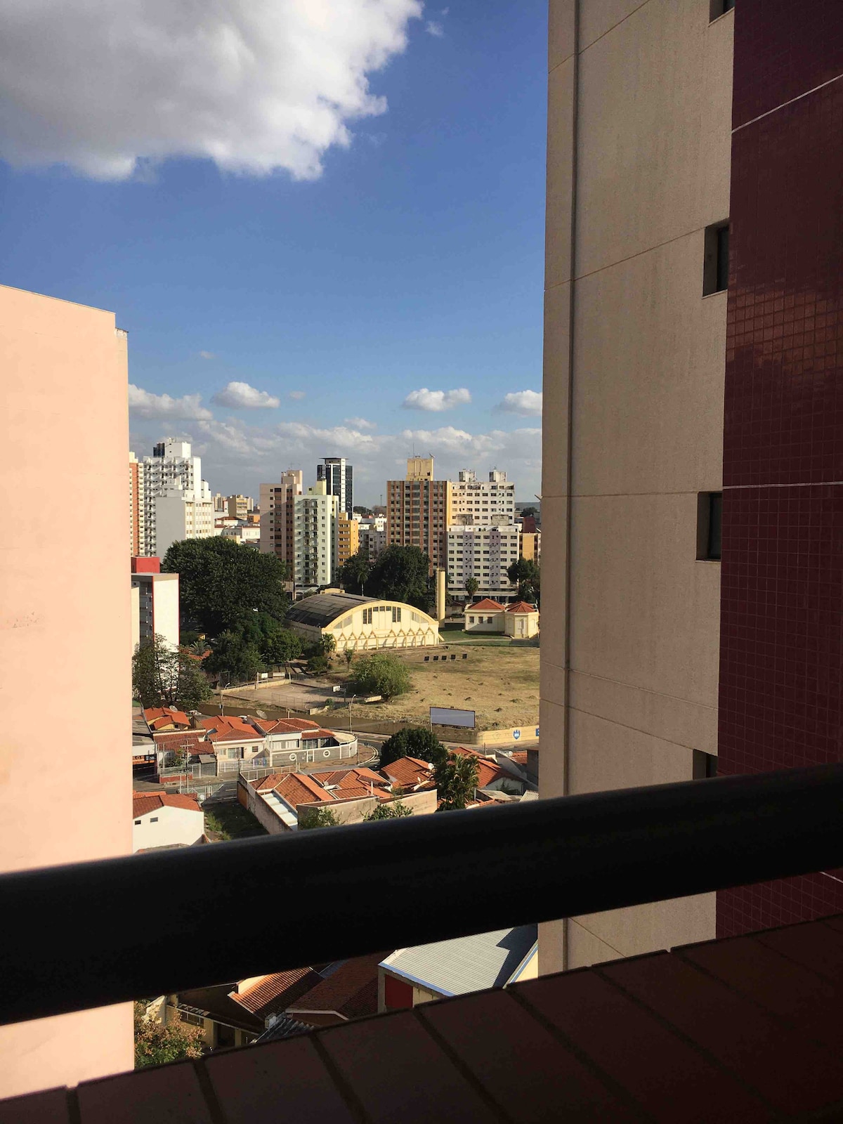 A view from the balcony showcases a mix of modern and traditional architecture, with residential buildings in the background and a green space in the foreground. The scene is brightened by a clear blue sky dotted with clouds.