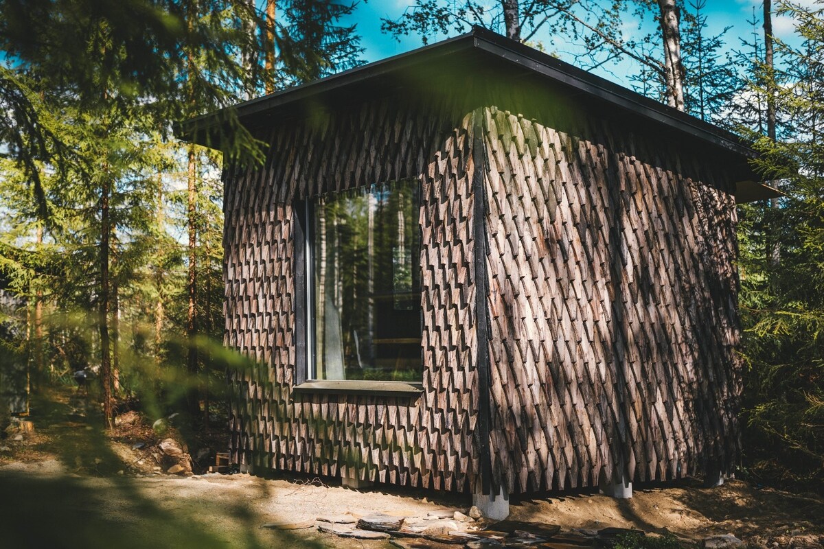 The cabin features a unique wooden exterior, with a textured pattern created by overlapping shingles. Large windows allow natural light to enter, providing views of the surrounding forest. The structure is set on a cleared area, nestled among tall trees.
