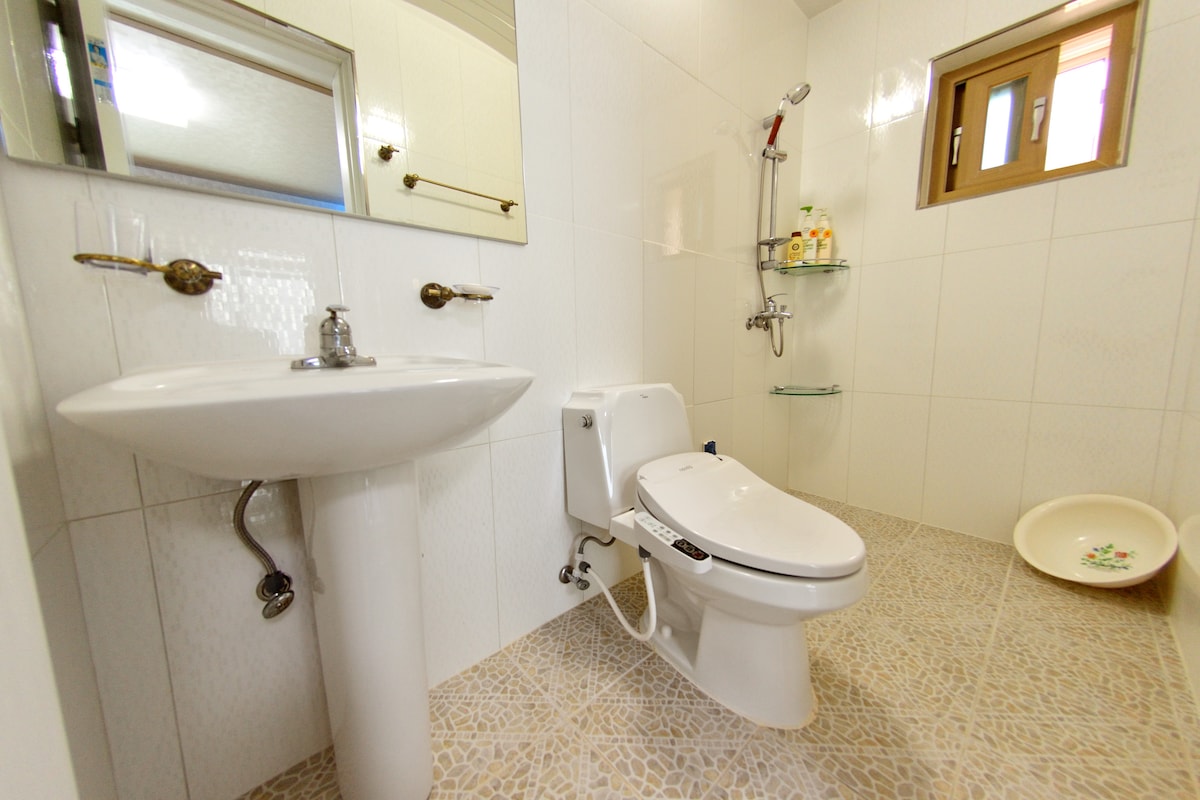 A clean and well-lit bathroom features white tiled walls and a large mirror above a modern sink. A toilet is positioned next to a shower area, which is equipped with shelves for toiletries. The floor is covered with a light-colored patterned tile.