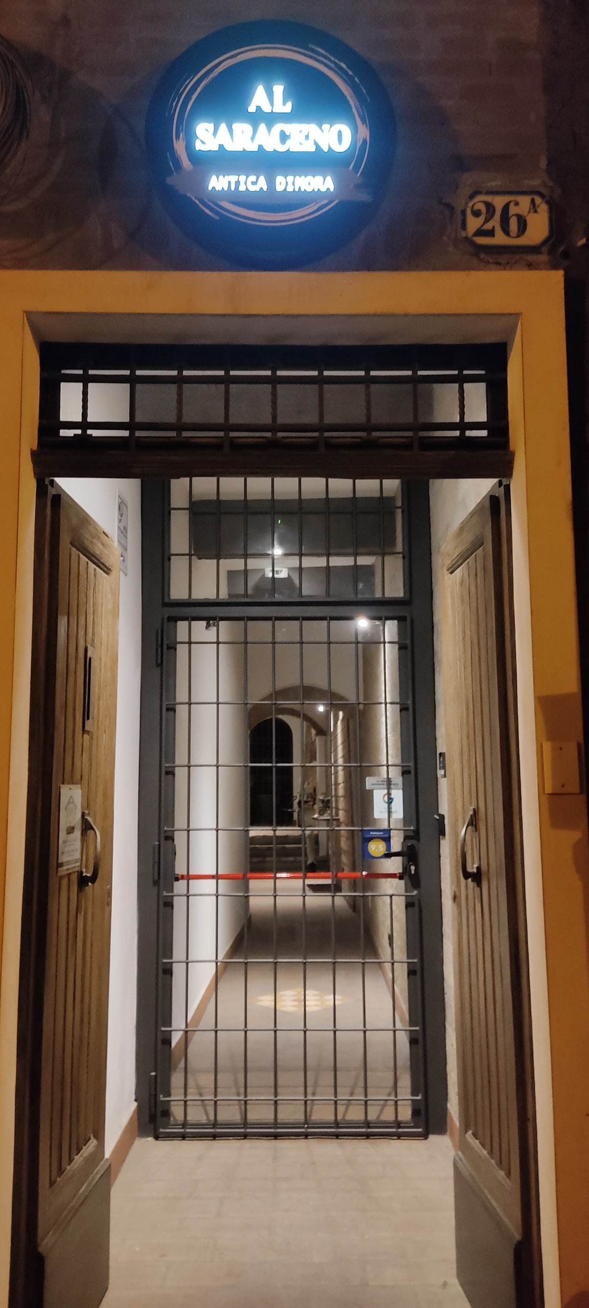 The entrance features double wooden doors with a metal grille, framed by a sign reading 'AL SARACENO.' A well-lit corridor can be seen beyond, showcasing a tiled floor that leads into the building.