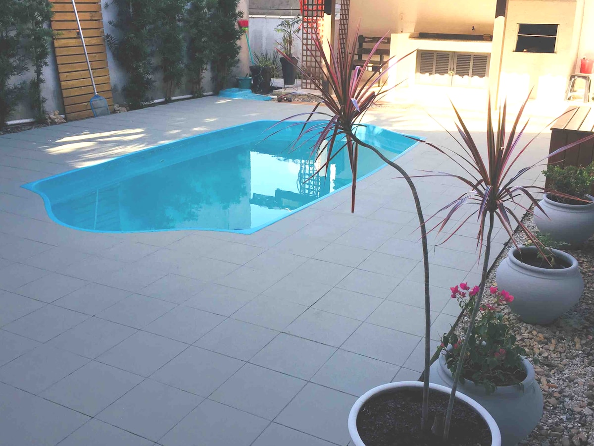 A private outdoor area features a shimmering pool surrounded by a tiled patio. Decorative plants in modern pots add natural elements to the space, and a barbecue area is visible in the background, enhancing the setting for gatherings.