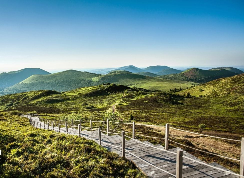 A scenic landscape features a winding wooden pathway lined with a rustic fence, leading through lush green hills. The panoramic view showcases rolling mountains under a clear blue sky, creating a serene and open atmosphere.