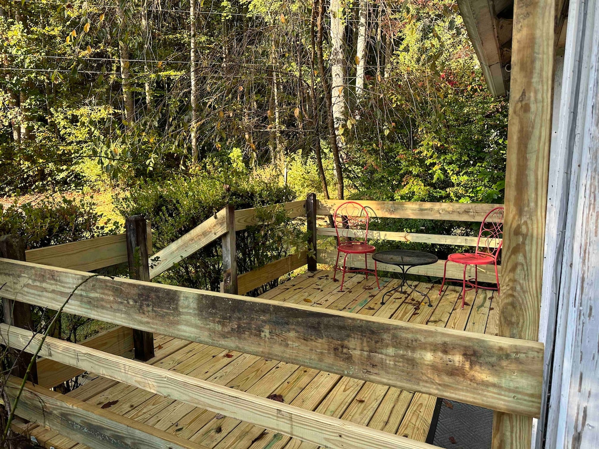 A wooden deck is surrounded by natural greenery, featuring two red chairs and a small table. The deck offers a peaceful outdoor space that blends with the surrounding landscape, highlighting the area's tranquility.