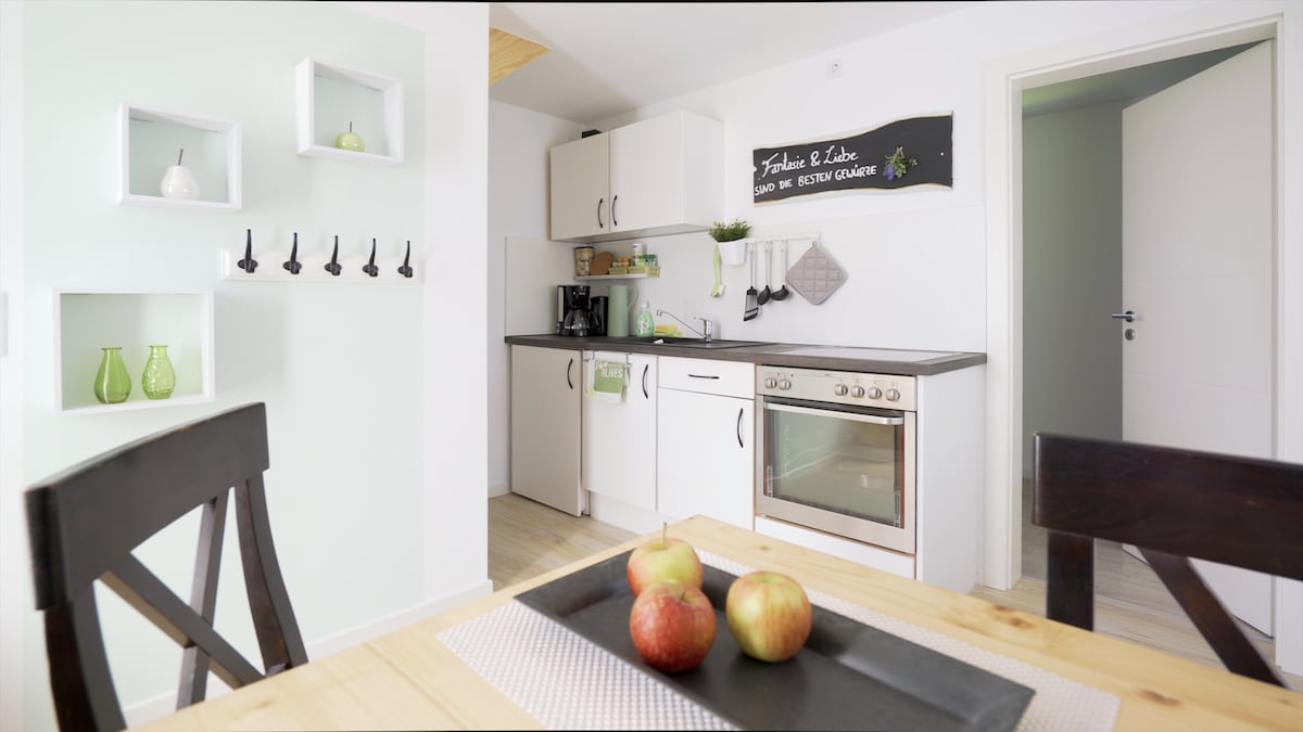 A modern kitchen is showcased with white cabinetry and a black countertop. Kitchen utensils hang on the wall, and decorative green vases add a touch of color. A dining table is set with a black placemat and three red apples as a centerpiece, enhancing the welcoming atmosphere.