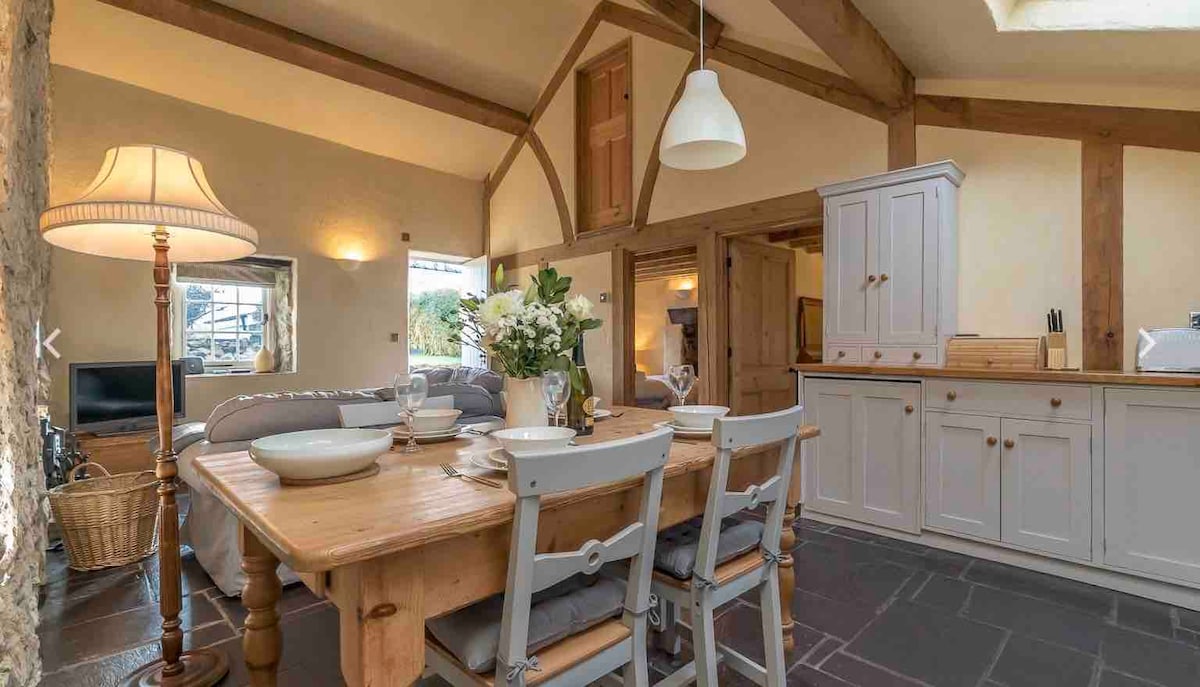 An open plan living space features a farmhouse-style dining table with grey chairs, set for a meal. A cozy sofa is visible in the background, with light streaming in through a window. The room highlights exposed wooden beams and slate tile flooring.