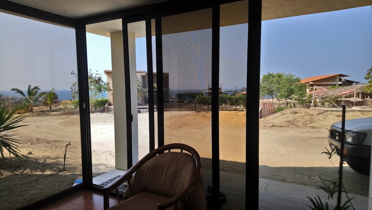 A serene outdoor view is framed by large glass windows, showcasing a spacious landscape with distant ocean views and nearby houses. A comfortable chair sits invitingly on the porch, surrounded by greenery and a clear path leading to the driveway.