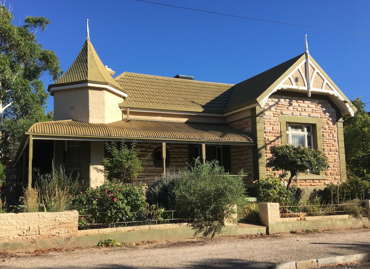 The heritage-listed building is characterized by its stone facade and unique architectural features, including a pointed turret and decorative gables. Surrounding greenery adds a natural touch, while a charming garden is visible in the foreground.