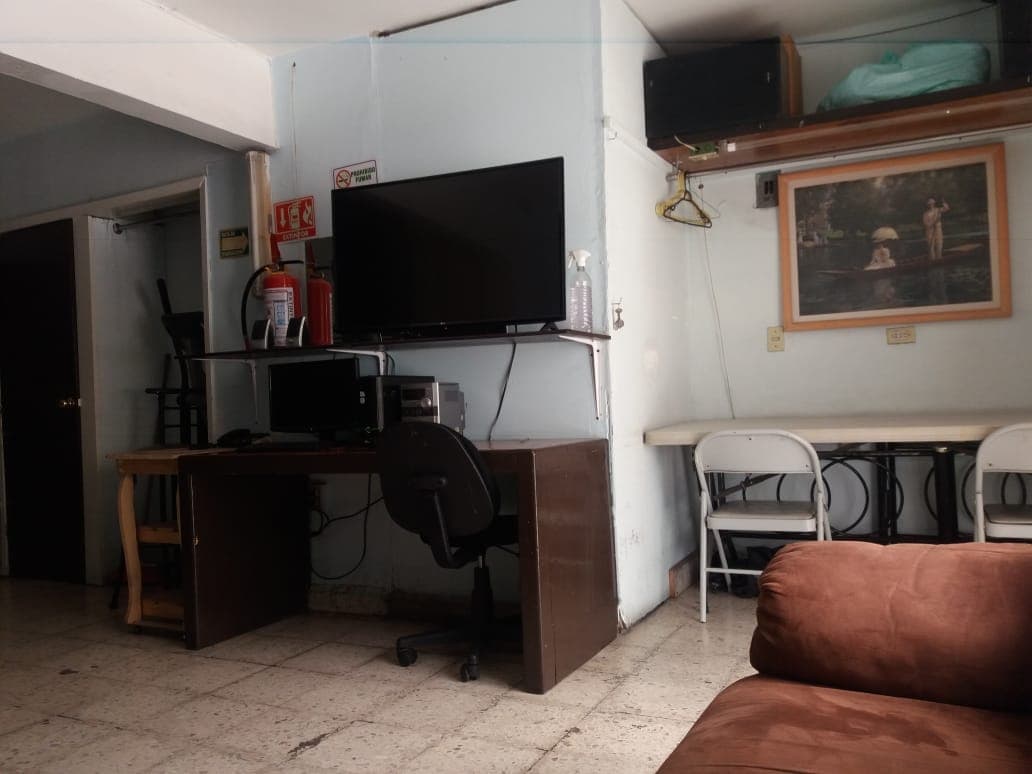 A functional living area is presented, featuring a wall-mounted television above a desk with a computer and office chair. To the side, a small dining table with two white chairs is visible. A comfortable brown couch invites relaxation in the space.