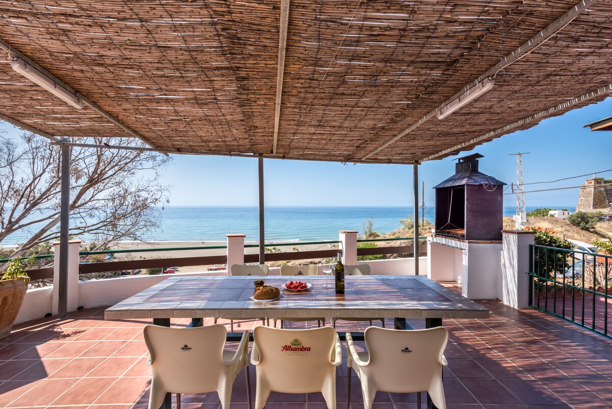 A spacious, covered patio is equipped with a large table and chairs, overlooking the serene sea. A barbecue area is visible on one side, and the open view of the water enhances the outdoor dining experience. Natural light floods the space through the open design.