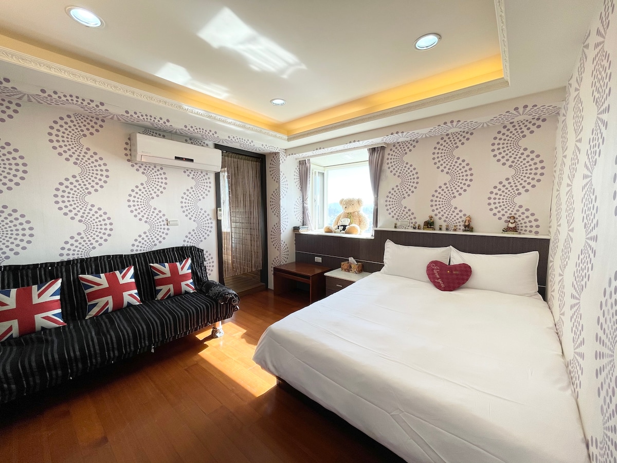 A spacious room features a cozy bed with crisp white linens and patterned wallpaper. A comfortable black sofa with Union Jack cushions adds character. Natural light illuminates the space through a large window, complemented by dark curtains. Decorative items are arranged on a wooden shelf.