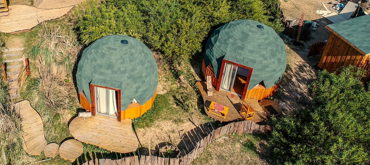 Two geodesic wooden domes are surrounded by natural greenery, featuring terraced decks with outdoor seating. The domes showcase large, circular windows and are designed with eco-sustainable materials. A winding pathway leads through the landscape, emphasizing the serene and tranquil setting.