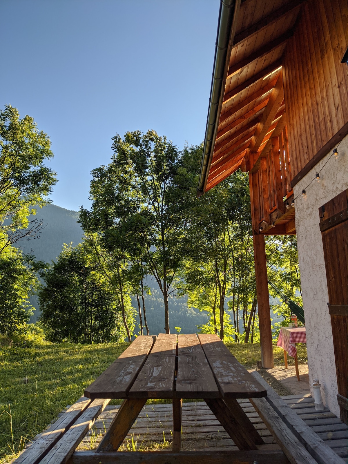 An outdoor area features a wooden picnic table situated under trees, providing a shaded spot for relaxation. The chalet's wooden structure extends into the scene, with mountains visible in the background. The sky is clear, enhancing the serene natural setting.