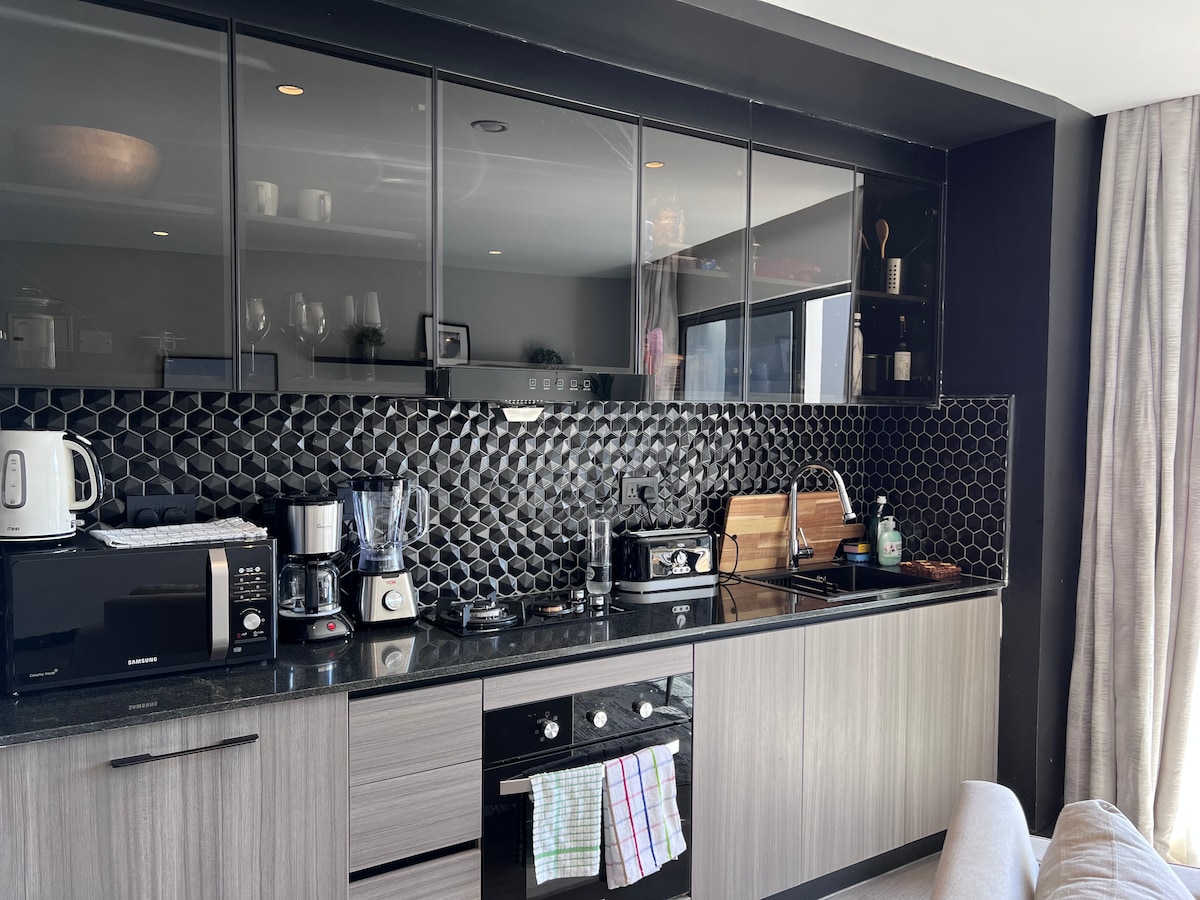 The kitchen area features modern cabinetry with a sleek black backsplash. Essential appliances, including a microwave, kettle, toaster, and coffee maker, are arranged for convenience. A gas stove with an oven, along with a well-organized countertop, ensures a functional cooking space.
