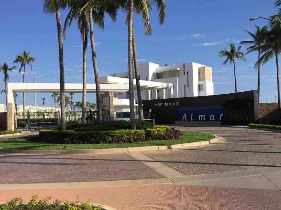 The entrance of Residencial Almar is depicted, featuring a modern architectural design and a welcoming sign. Tall palm trees line the driveway, and a landscaped circular area includes vibrant greenery. A paved path leads towards the secure entrance, framed by blue skies.