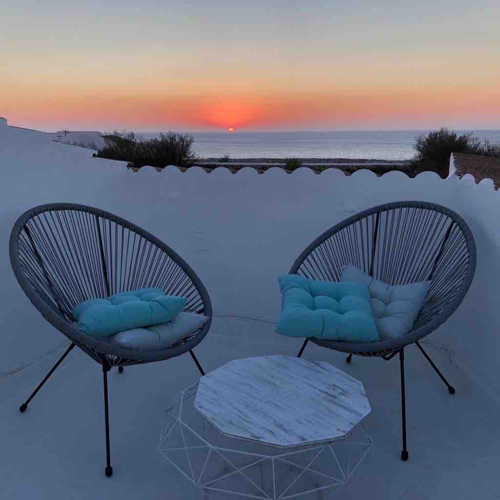 Two modern, circular chairs with light blue cushions are situated on a terrace, facing towards a serene sunset over the ocean. A white, geometric table stands between the chairs, providing a subtle contrast to the soft colors of the evening sky.