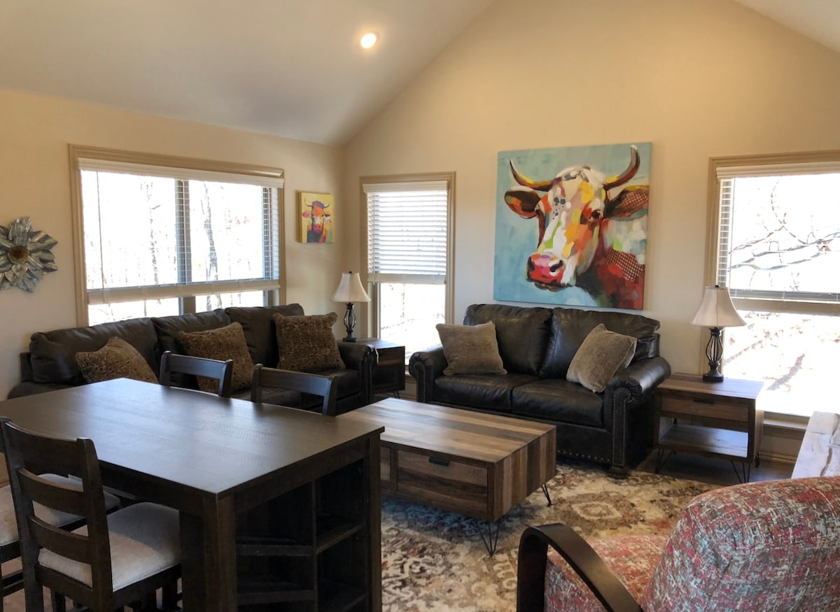 A spacious living area features two leather sofas and matching armchairs arranged around a wooden coffee table. Large windows allow natural light to fill the room, while a vibrant painting of a cow adds a touch of color to the neutral walls.