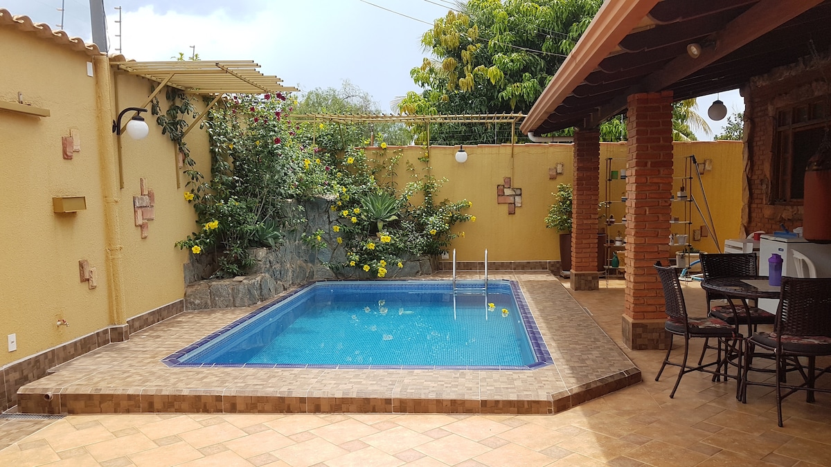 A private outdoor area features a rectangular pool surrounded by a tiled patio and vibrant greenery. Flowering plants cascade from walls, adding color. A seating area with chairs and a table is positioned beside the pool, with a covered section providing shade.